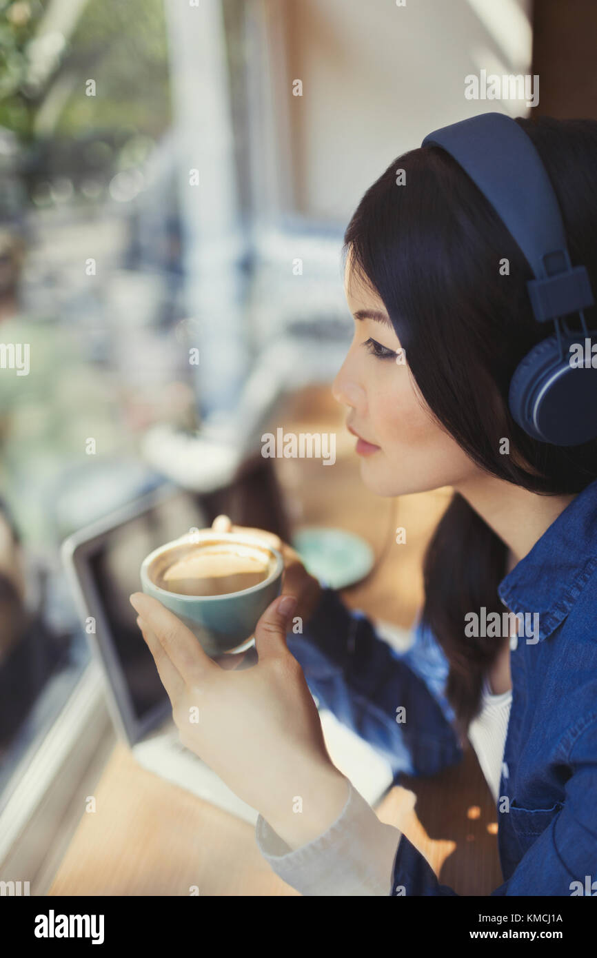 Pensive young woman drinking coffee, listening to music with headphones at cafe window Stock Photo