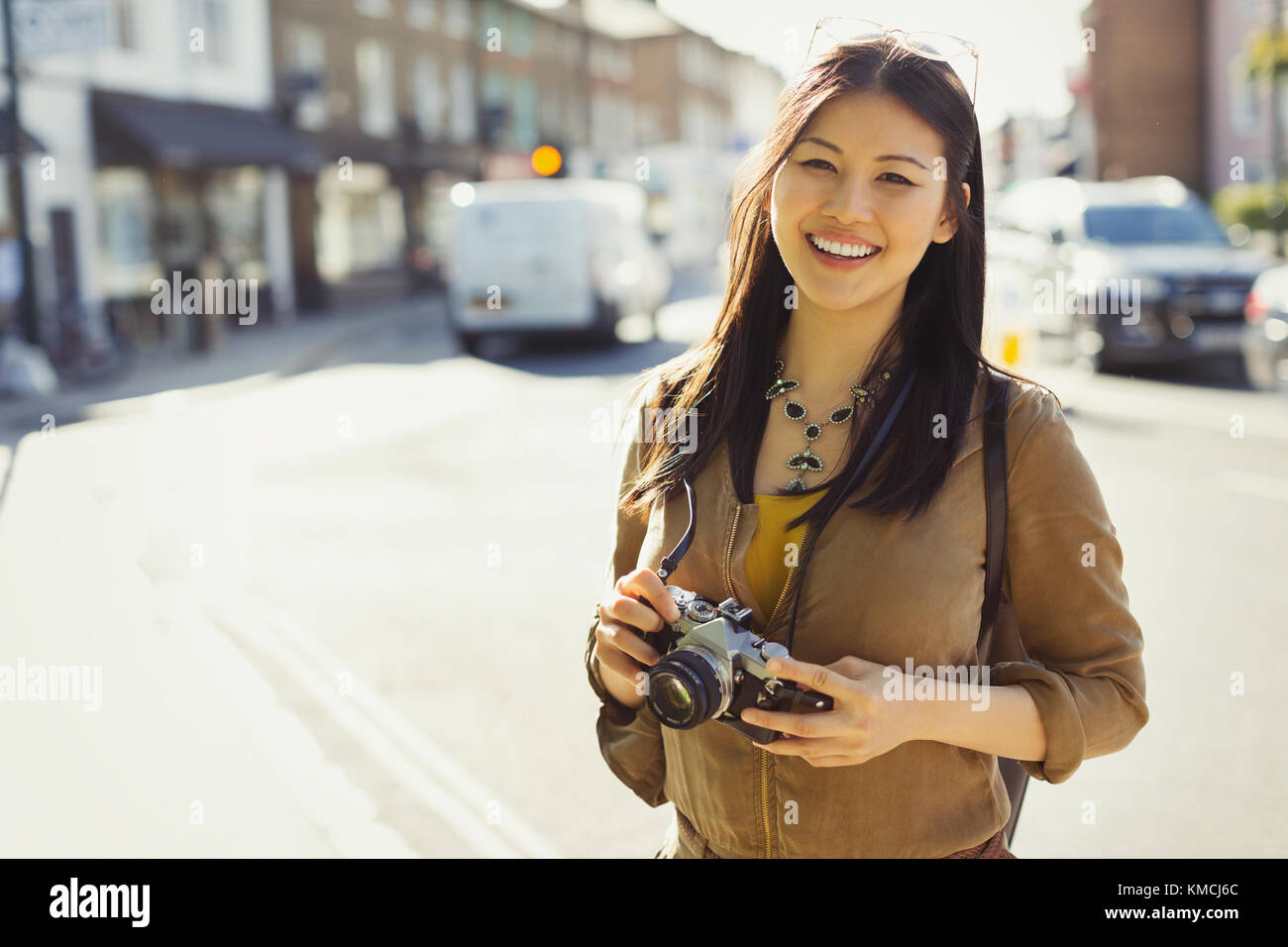 Portrait smiling, confident young female tourist with camera on sunny urban street Stock Photo