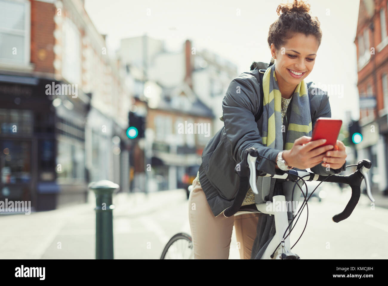 Smiling young woman commuting with bicycle, texting with cell phone on sunny urban street Stock Photo