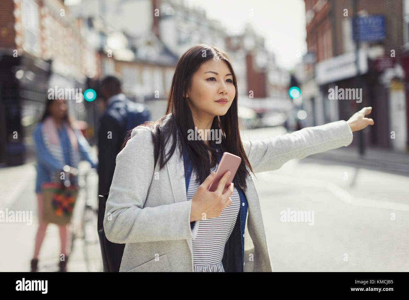 Young businesswoman with cell phone hailing taxi on sunny urban street Stock Photo