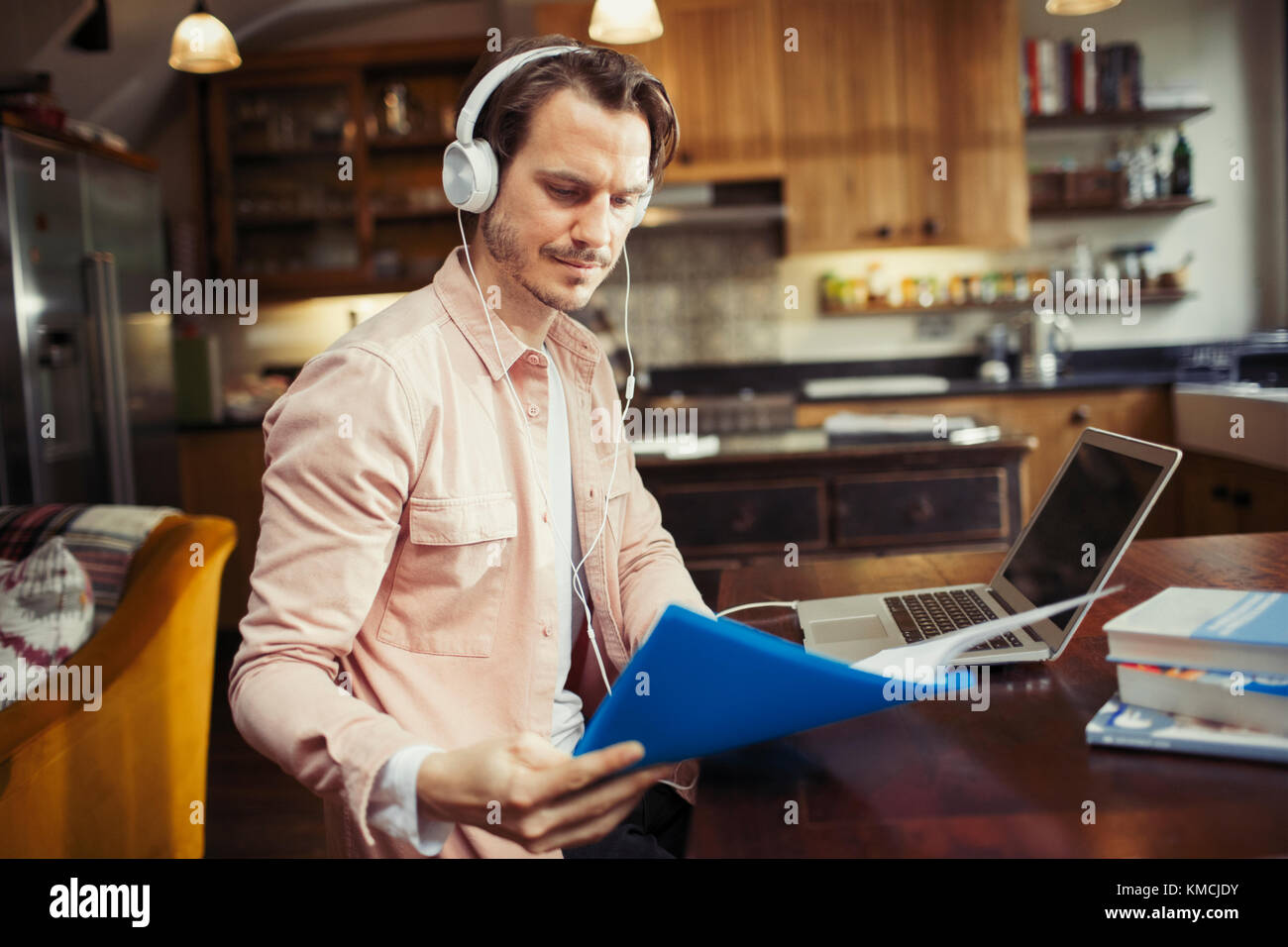 Man with headphones working at laptop, reading paperwork in kitchen Stock Photo