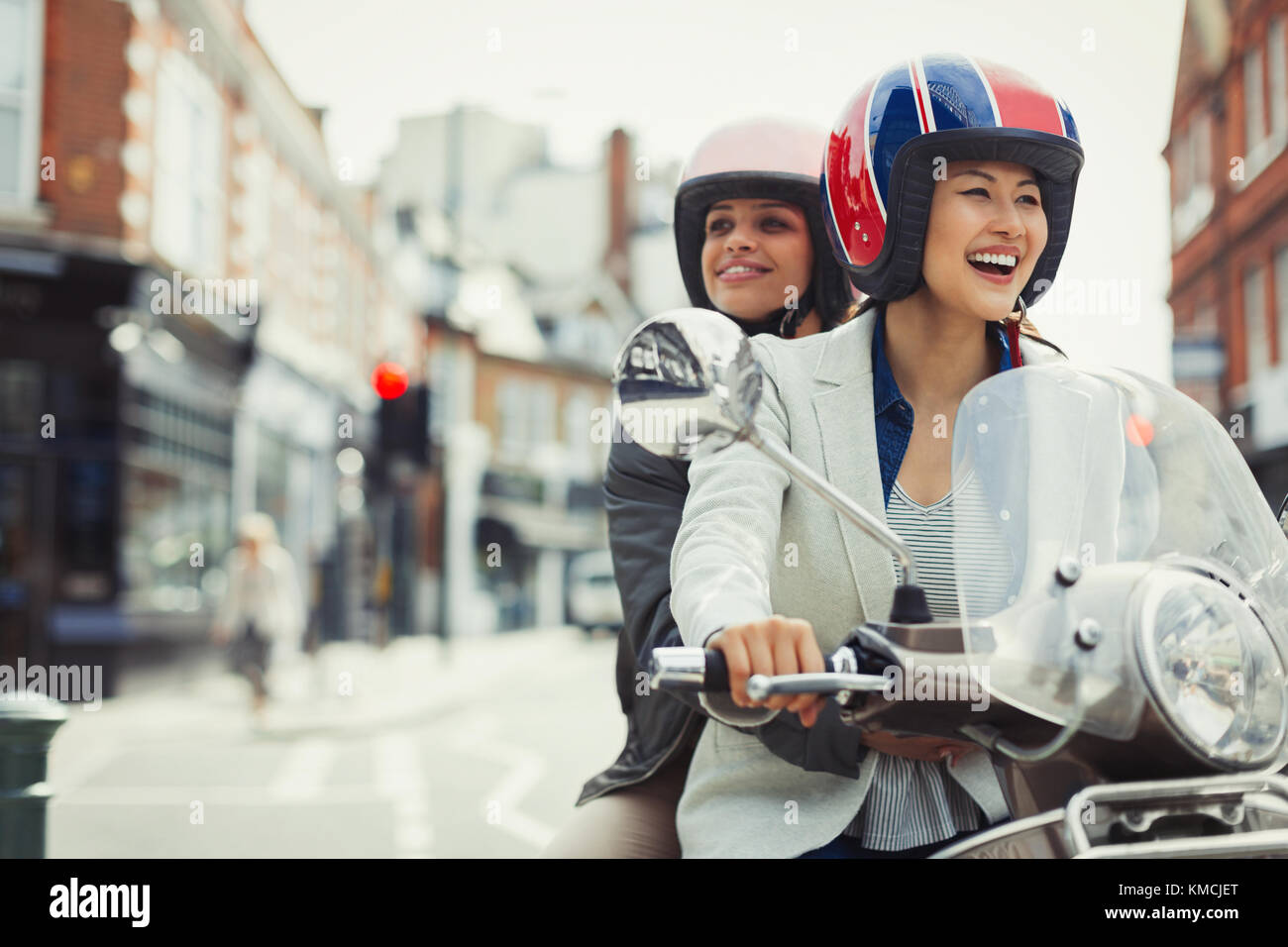 Smiling young women friends wearing helmets and riding motor scooter on urban street Stock Photo