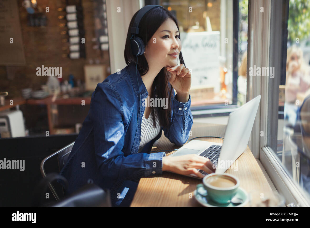 Pensive young woman listening to music with headphones at laptop and drinking coffee in cafe window Stock Photo