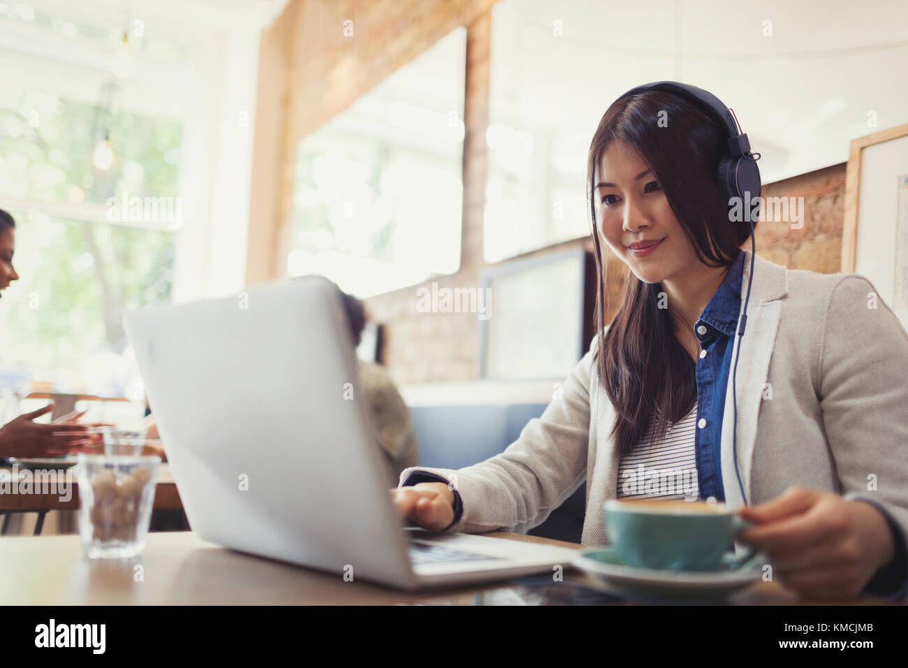 Smiling young woman listening to music with headphones at laptop and drinking coffee in cafe Stock Photo