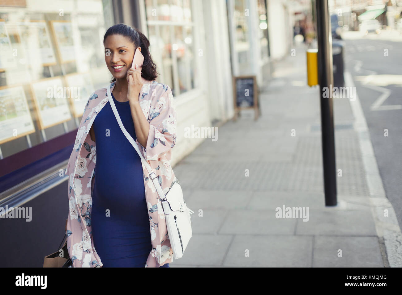 Smiling pregnant woman talking on cell phone, walking along urban storefront Stock Photo