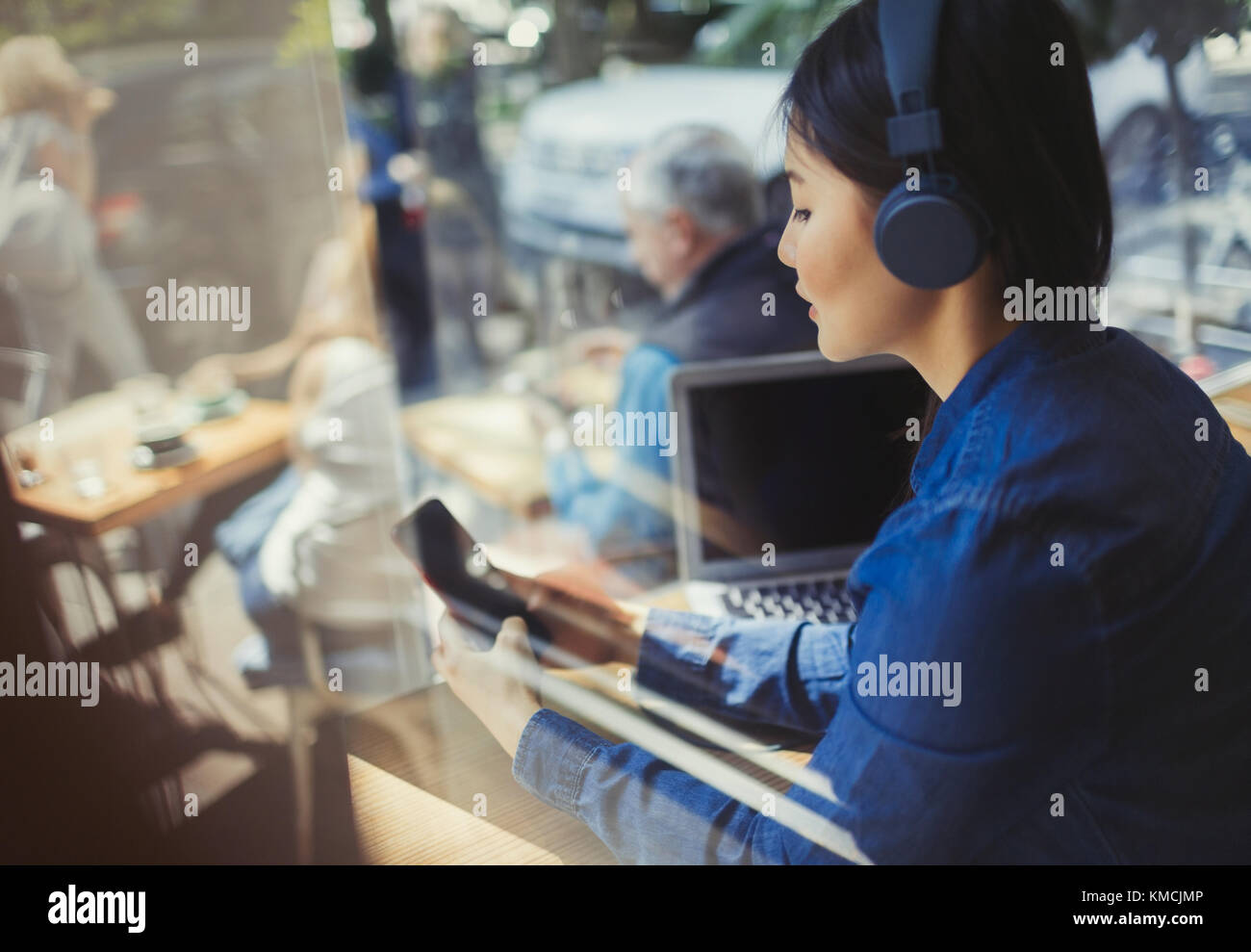 Young woman listening to music with headphones, texting with cell phone at cafe window Stock Photo