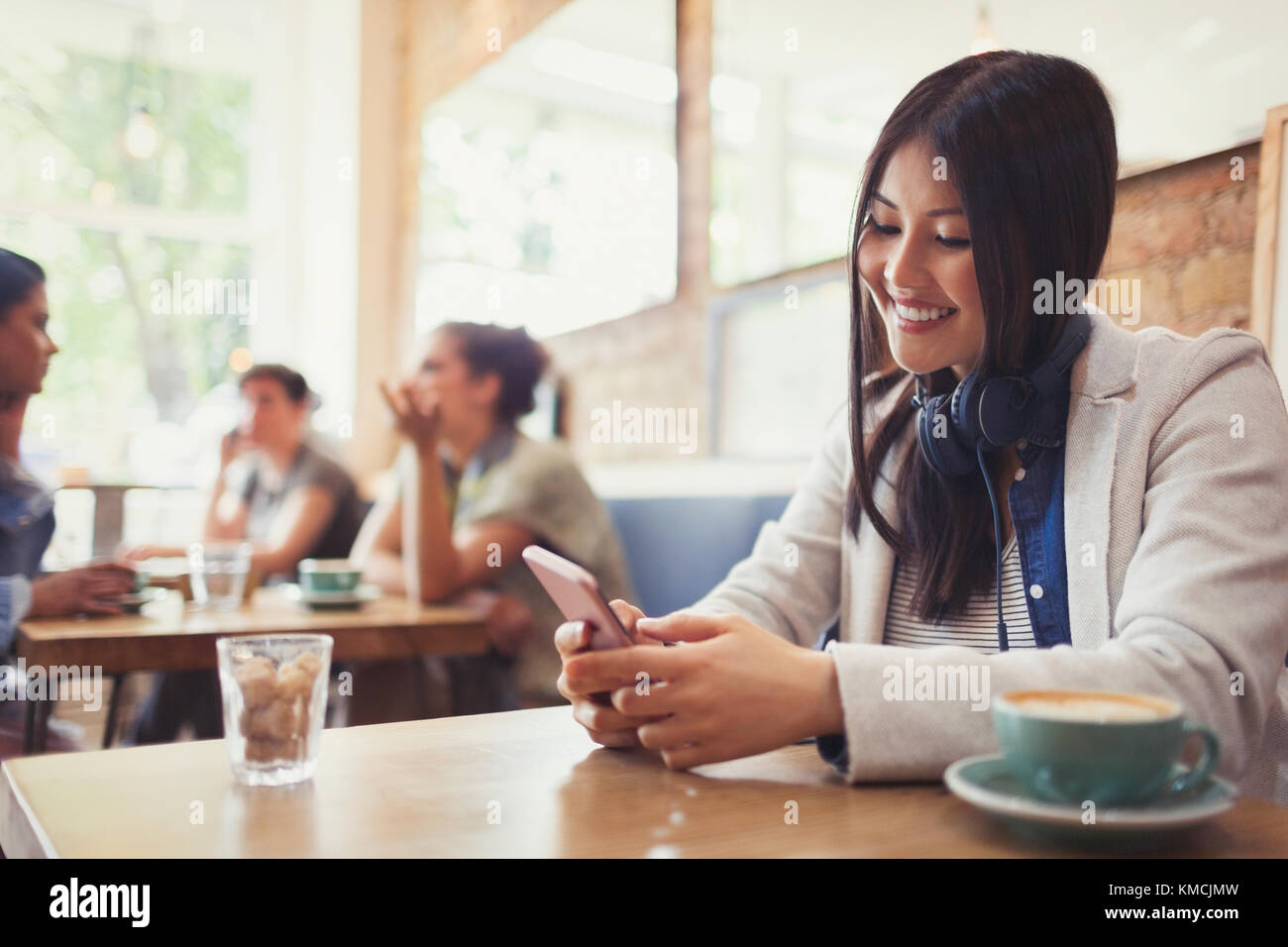 Smiling young woman with headphones texting with cell phone and drinking coffee at cafe table Stock Photo
