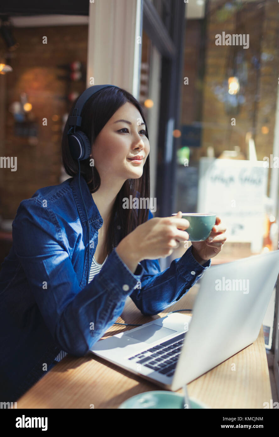 Pensive young woman listening to music with headphones and drinking coffee at laptop in cafe window Stock Photo