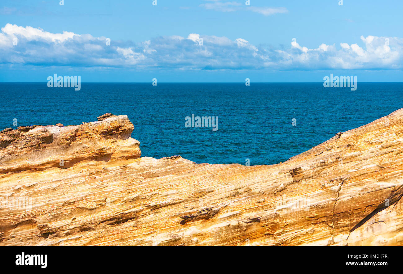 Sandstone contrasts with the blue waters of the Pacific ocean.  A line of clouds hovers over the horizon in the blue sky Stock Photo