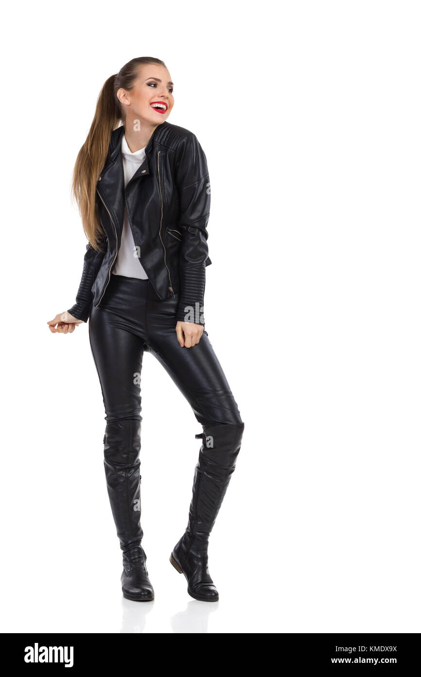 Smiling young woman in black leather trousers, jacket and boots posing and looking away. Full length studio shot isolated on white. Stock Photo