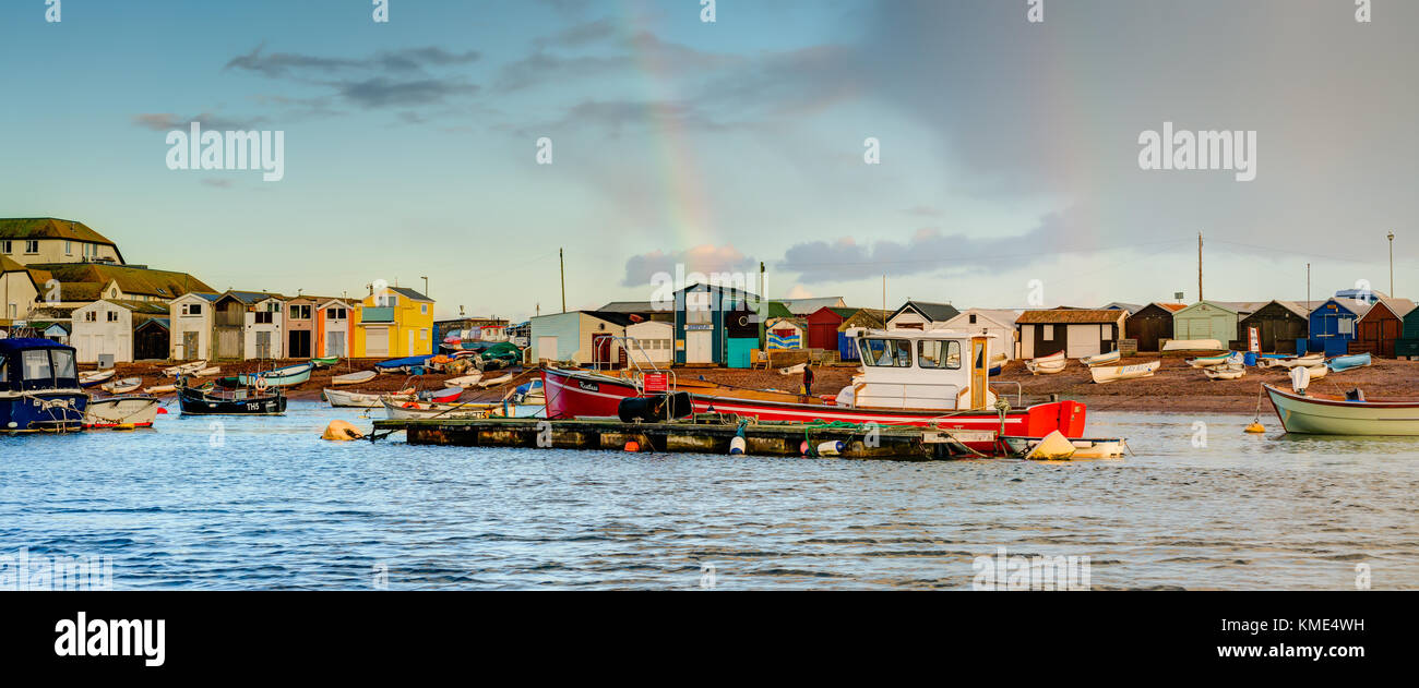 A colourful panoramic of 'life on the estuary' showing moored boats and a variety of beach huts and buildings on the shoreline, a double rainbow too. Stock Photo