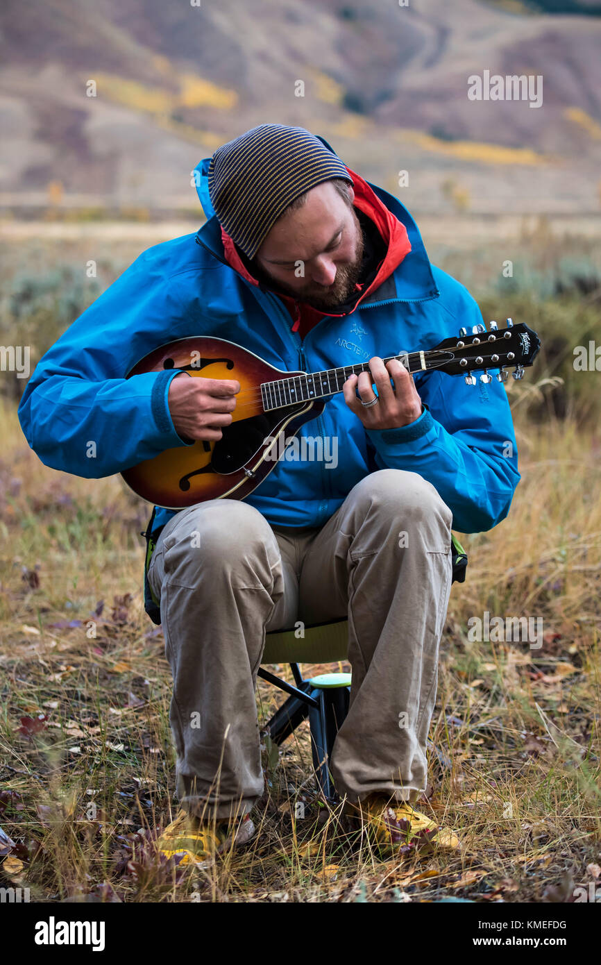 Male camper in jacket and knit hat playing mandolin while sitting,Jackson,Wyoming,USA Stock Photo