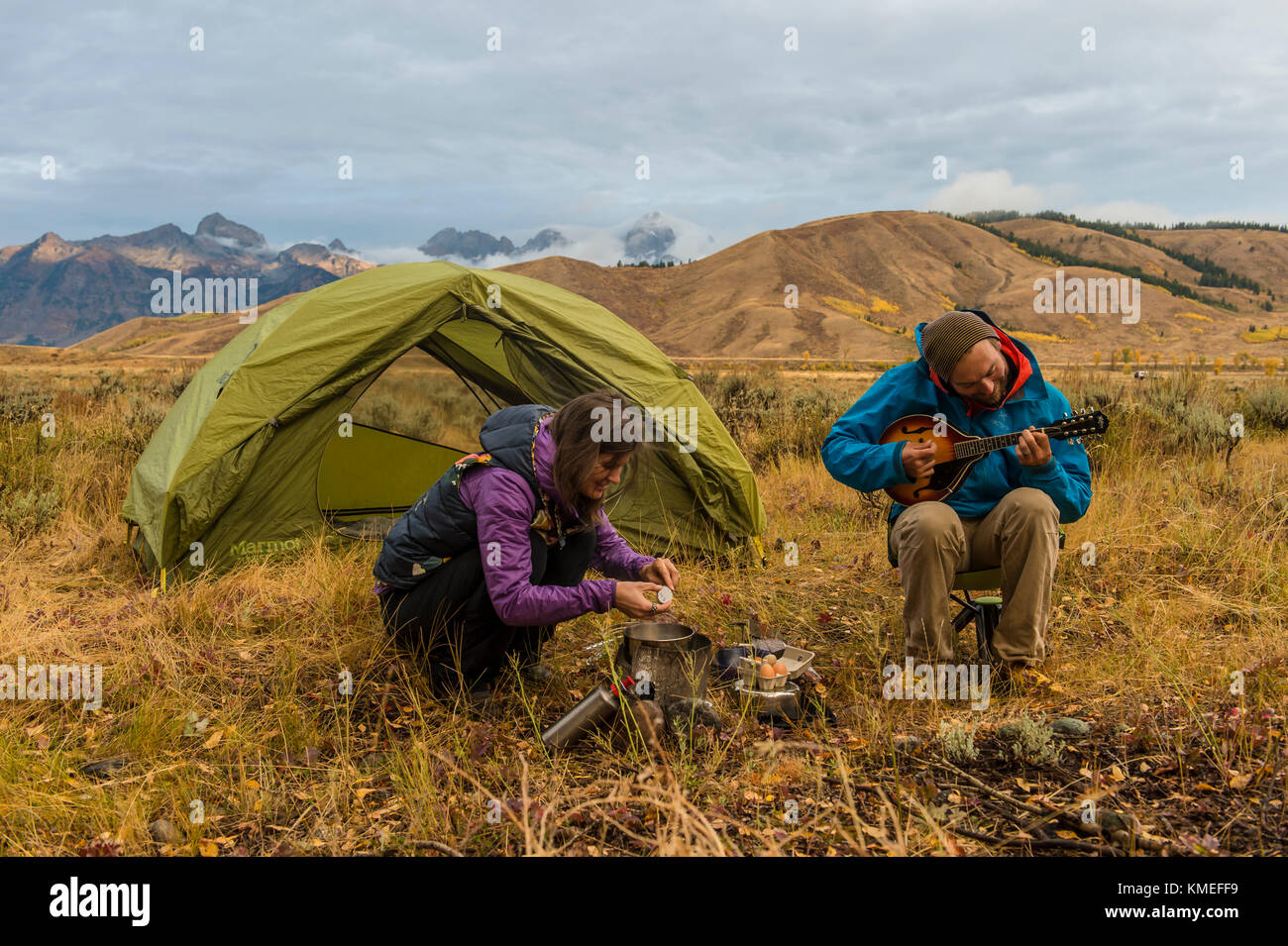 Female camper cooking breakfast on camping stove and male camper sitting and playing mandolin in front of tent,Jackson,Wyoming,USA Stock Photo