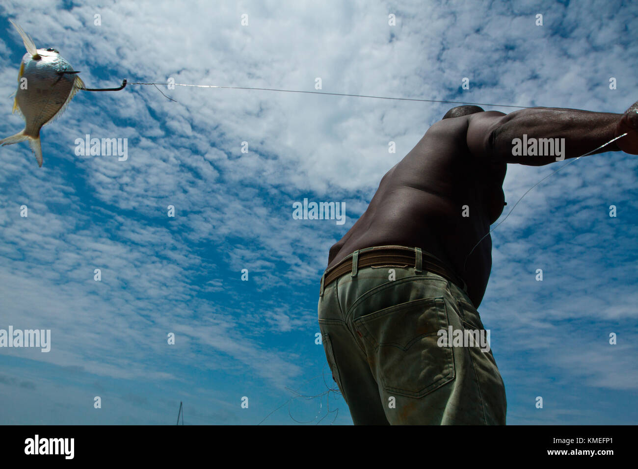 View from below of shirtless man casting fishing line with baitfish attached, Isla Marisol, Belize Stock Photo