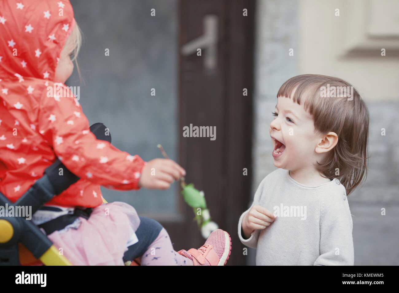Happy kid with long blond hair playing with a little girl sitting in a baby stroller. Stock Photo