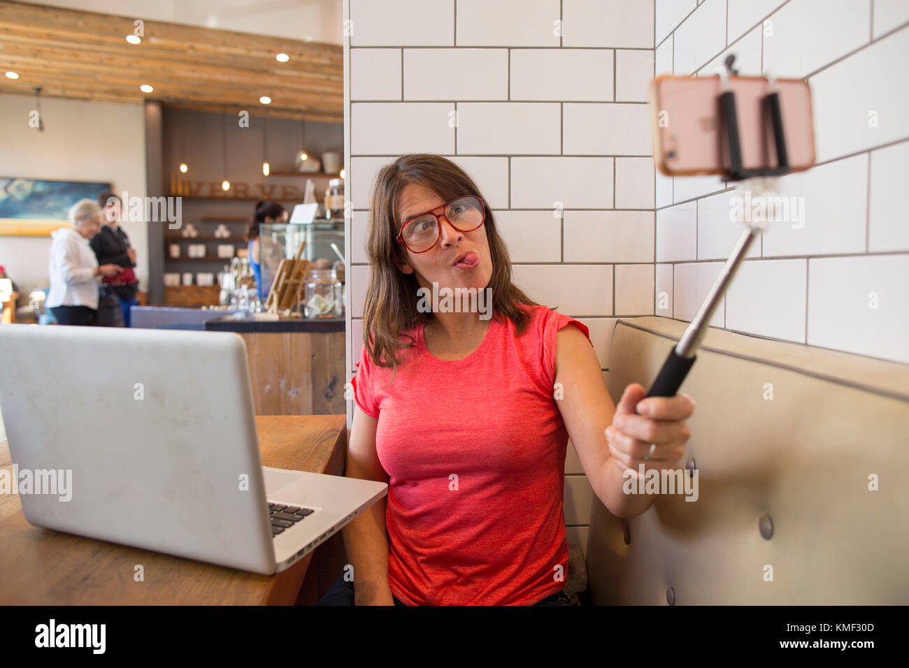 Woman making funny face while taking selfie in cafe with selfie stick,Santa Cruz,California,USA Stock Photo