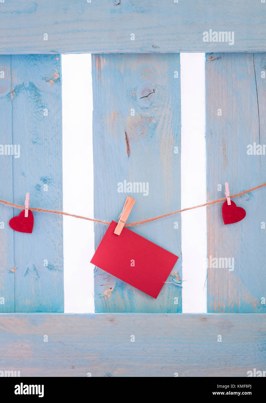 Unwritten red paper note surrounded by red hearts, tied to a linen string with wooden clips, on a blue fence. Stock Photo