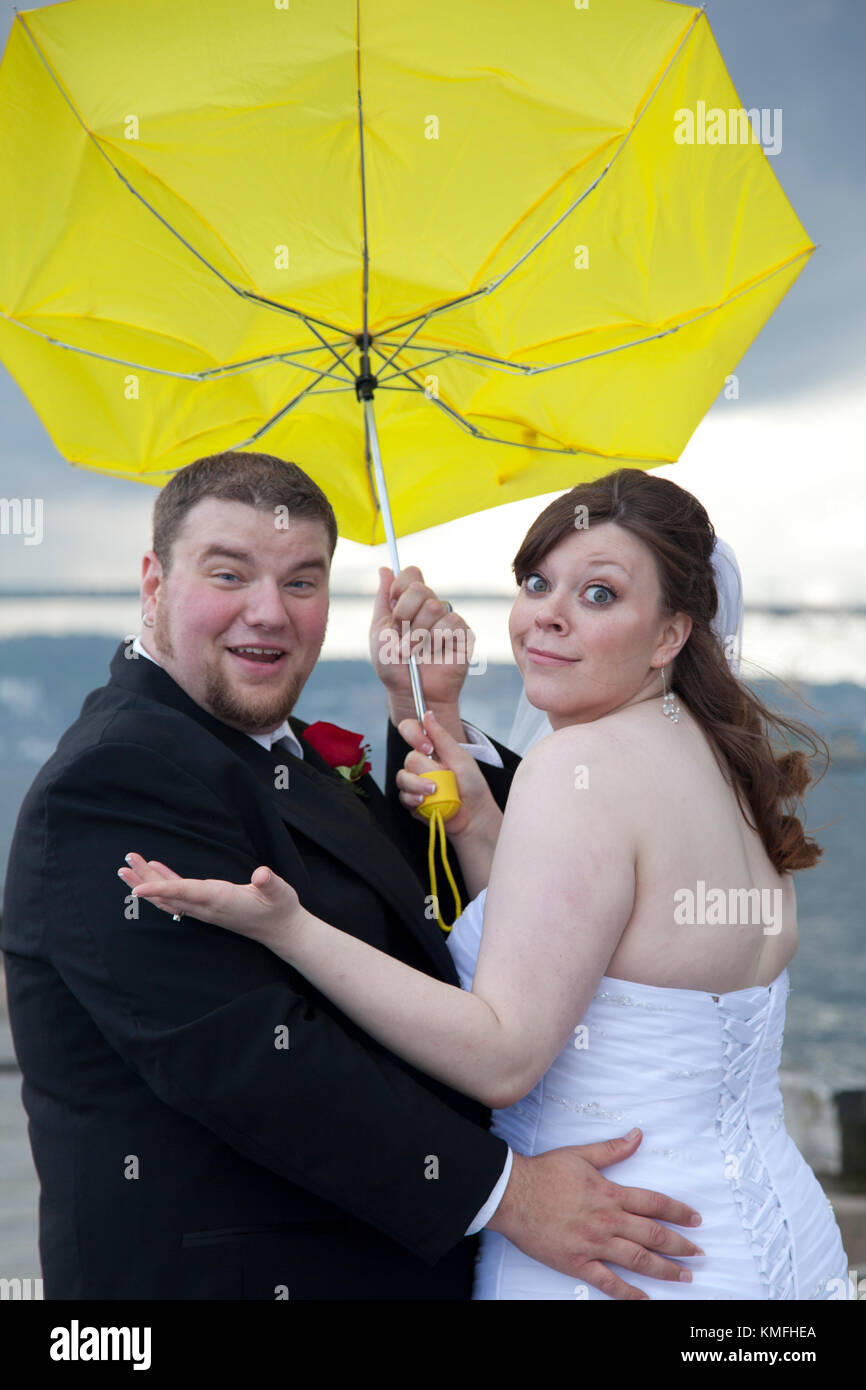 a newlywed couple shrugs as their umbrella turns inside out on their rainy wedding day Stock Photo