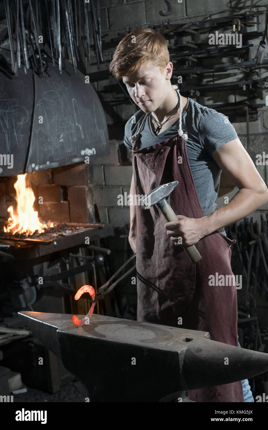 Apprentice blacksmith hammering red hot horseshoe on anvil at workshop Stock Photo