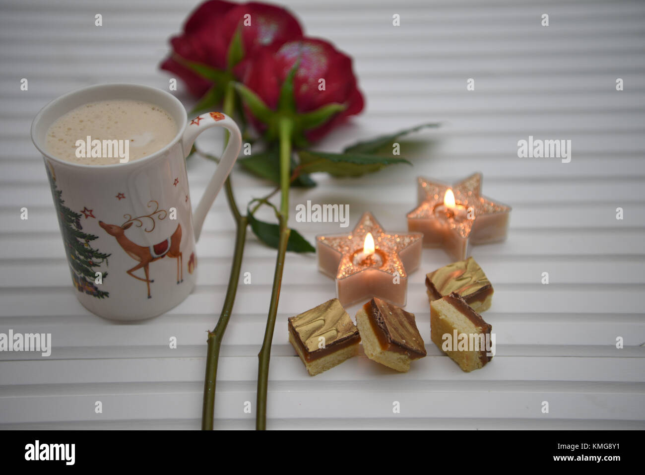 Christmas photography image with frothy hot drink in reindeer print mug with warm candles and luxury chocolate food on white wood background Stock Photo