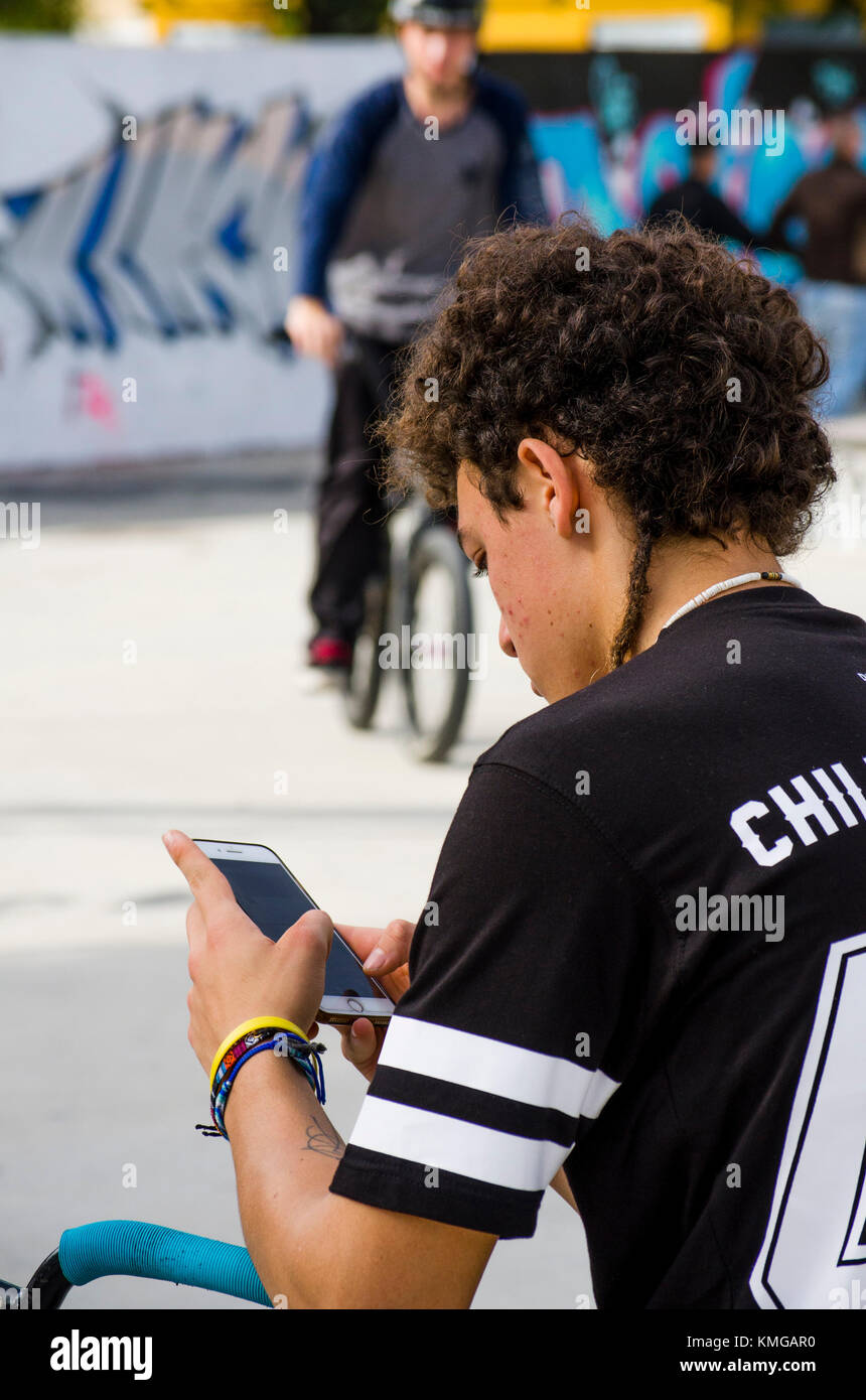 Teenage boy, 16-17 years texting on iPhone at skate park, Spain. Stock Photo