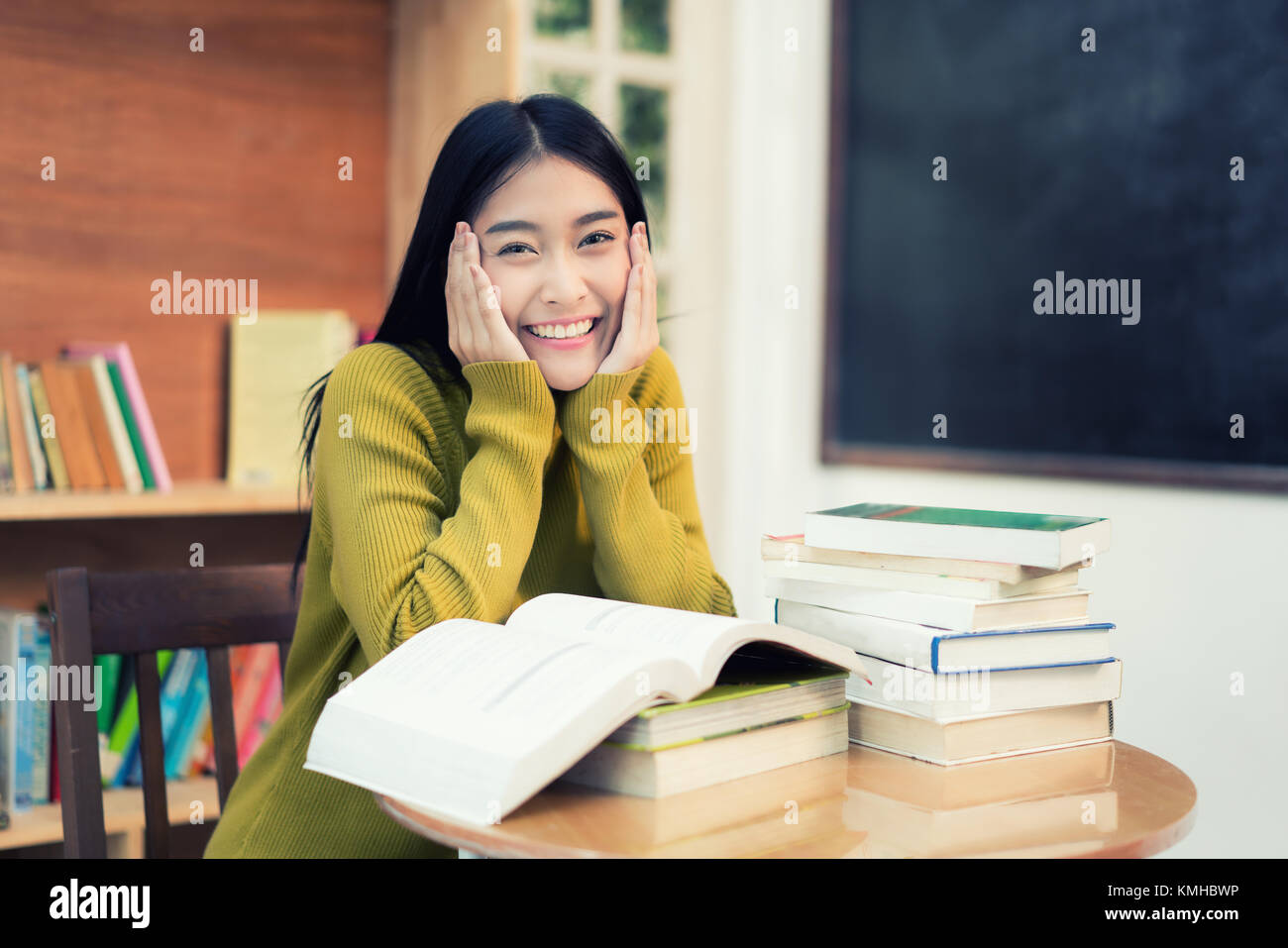 Cheerful female Asian student reading book during break between lesson near bookshelves in modern interior library of university. Stock Photo