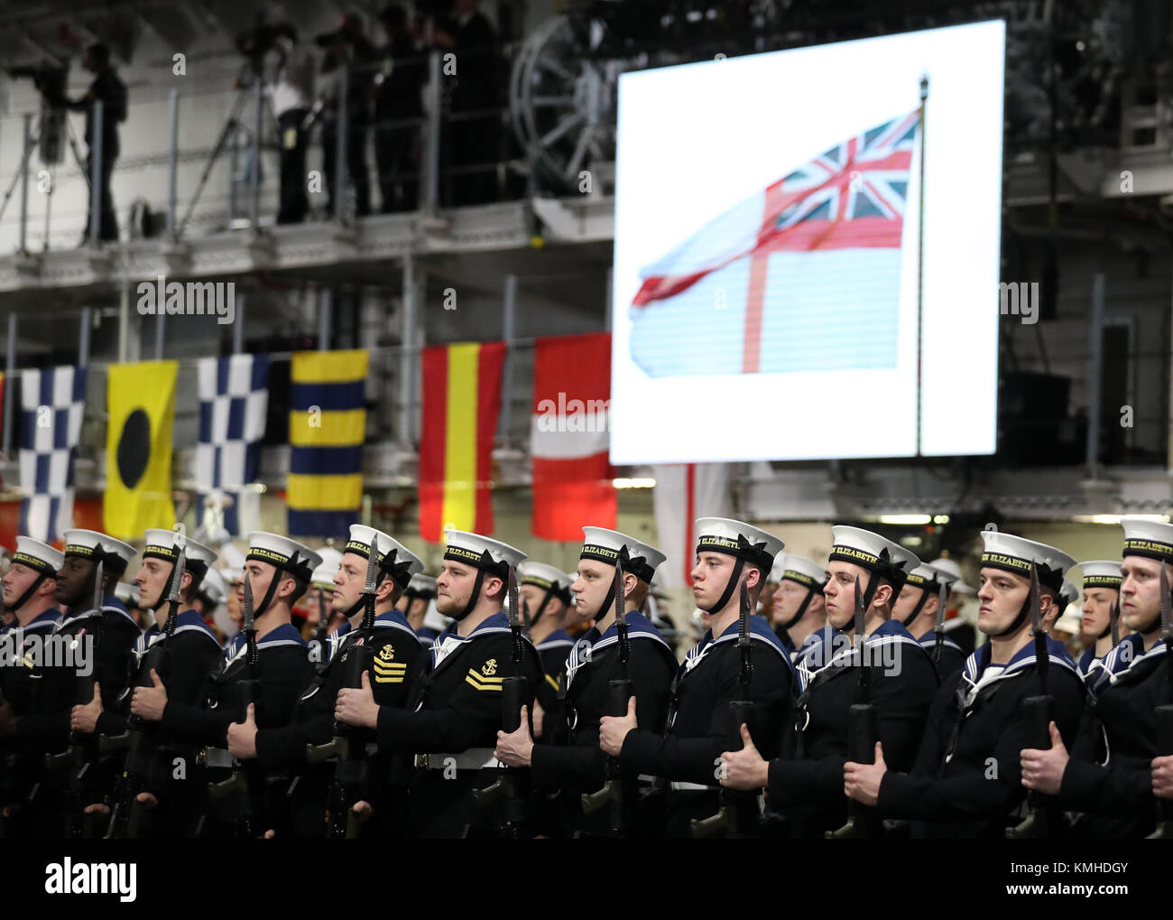Royal Navy ratings in front a screen showing the White Ensign being hoisted, in the hanger of HMS Queen Elizabeth during her commissioning ceremony in Portsmouth. Stock Photo