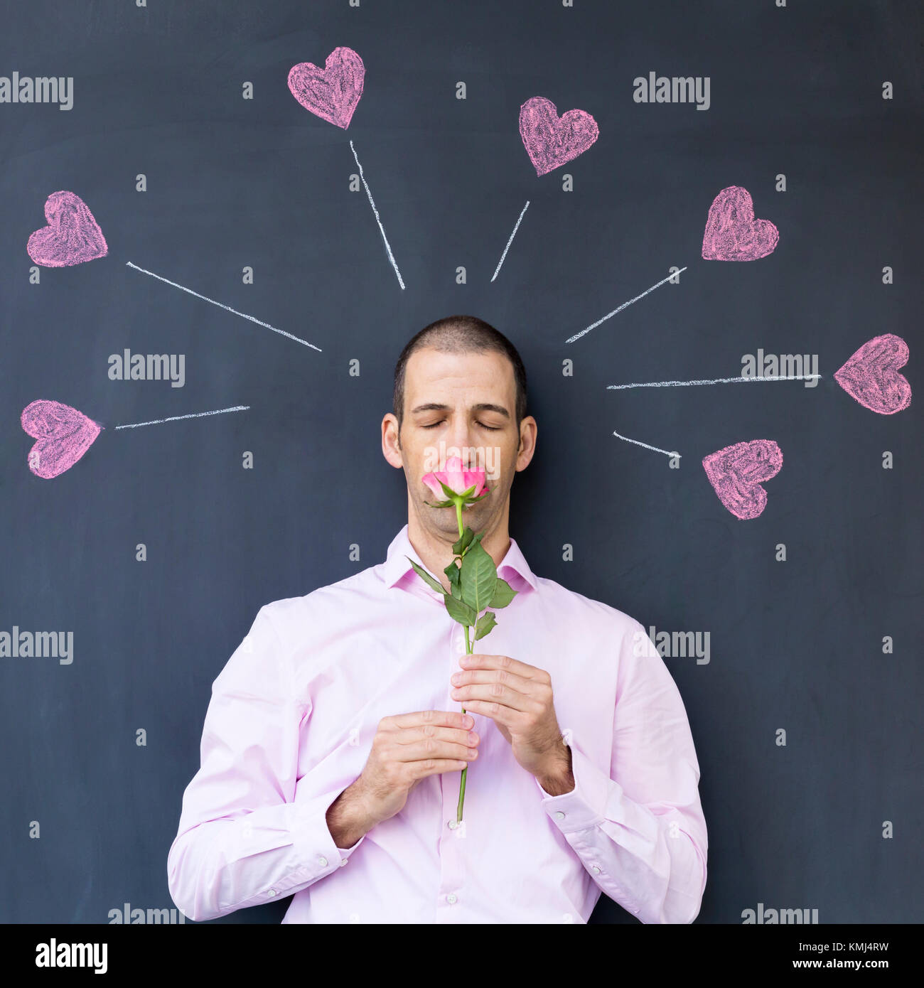 Single adult white man wearing a pink shirt standing in front of a blackboard with painted hearts holding a rose. Concept of crazy love. Stock Photo