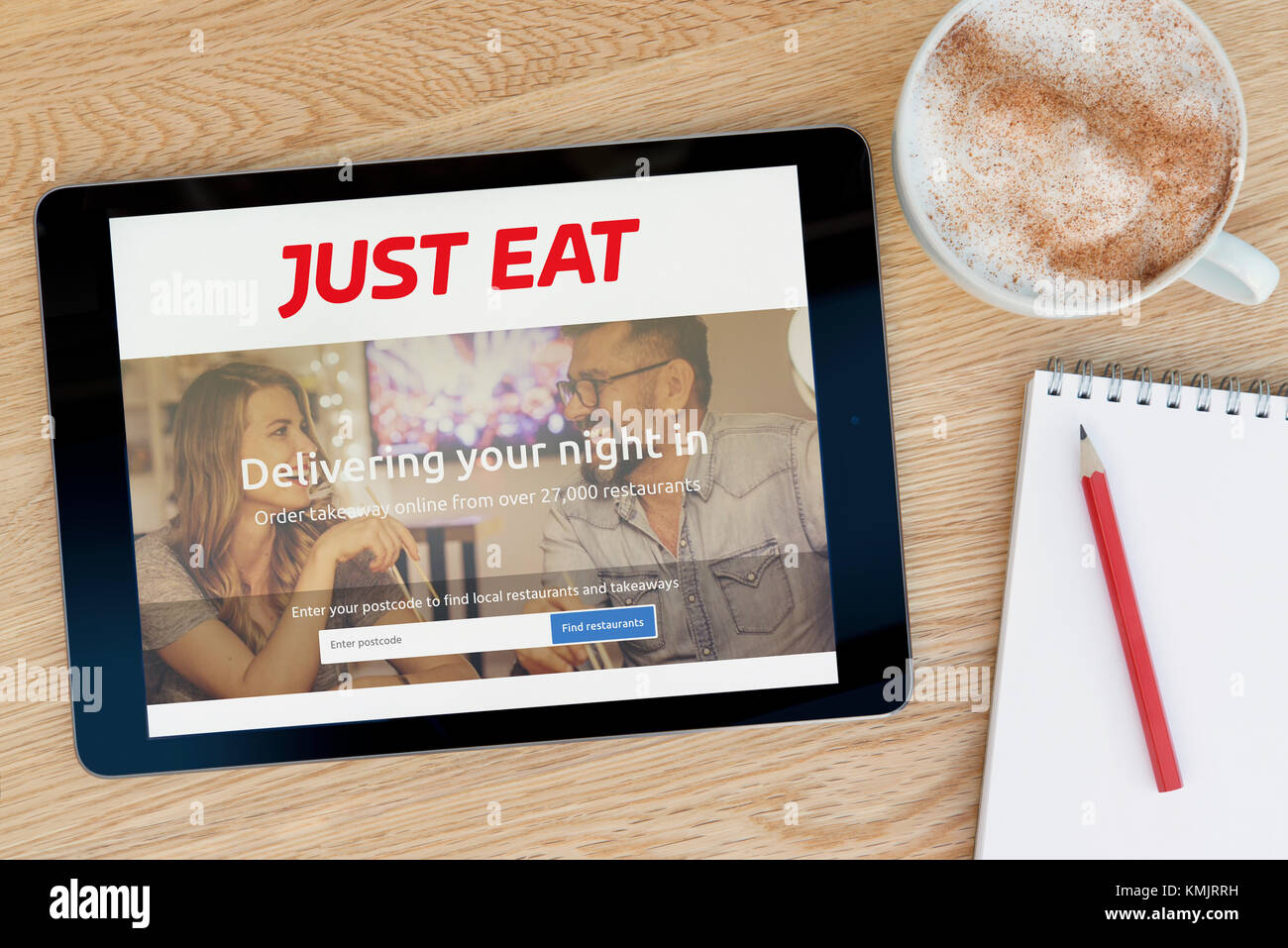 The Just Eat website features on an iPad tablet device which rests on a wooden table beside a notepad and pencil and a cup of coffee (Editorial only) Stock Photo