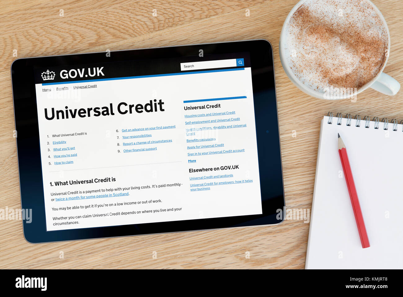 The Universal Credit part of the gov.uk website on an iPad tablet device rests on wooden table with notepad & pencil & cup of coffee -Editorial only Stock Photo