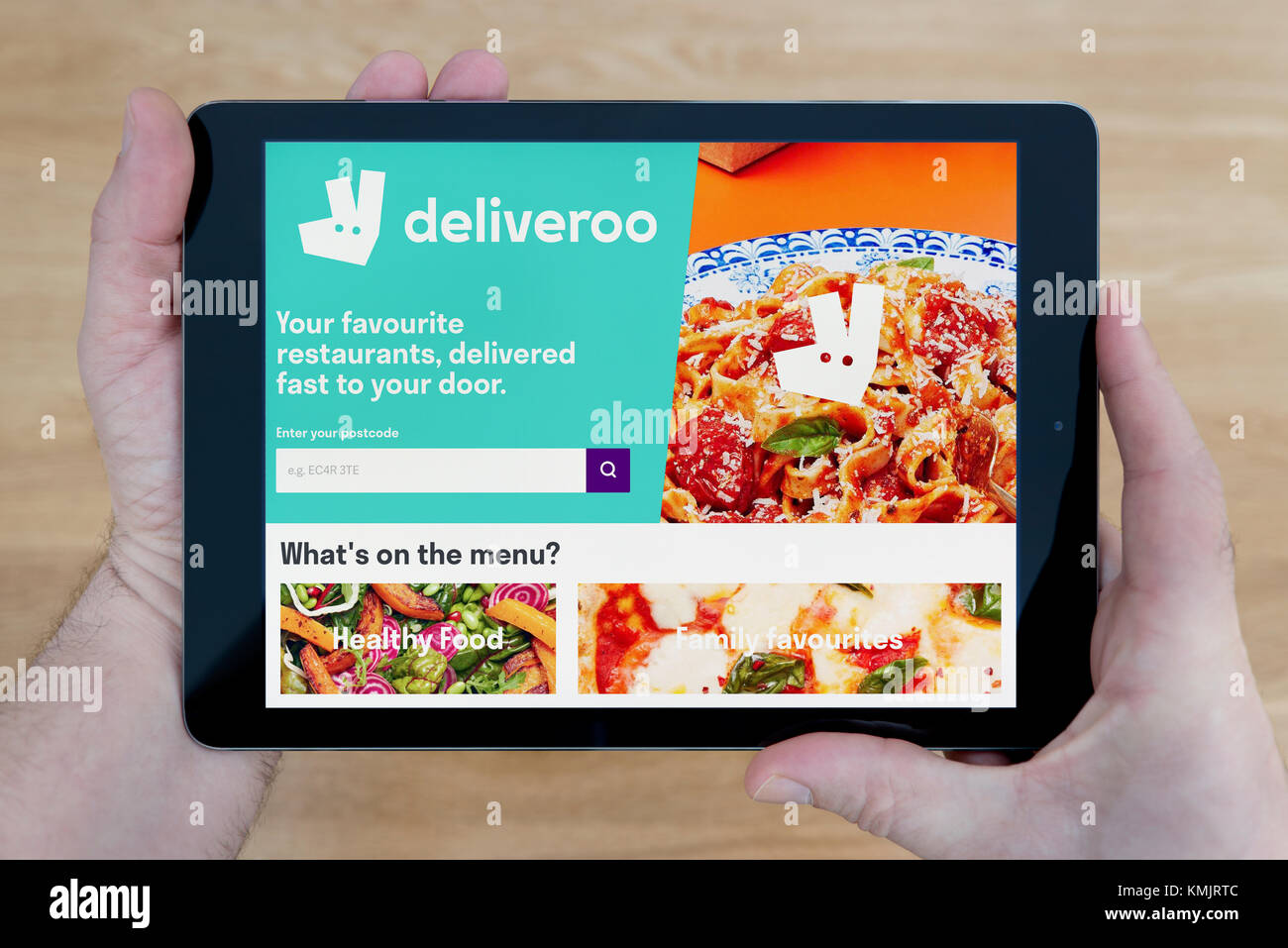 A man looks at the Deliveroo website on his iPad tablet device, shot against a wooden table top background (Editorial use only) Stock Photo