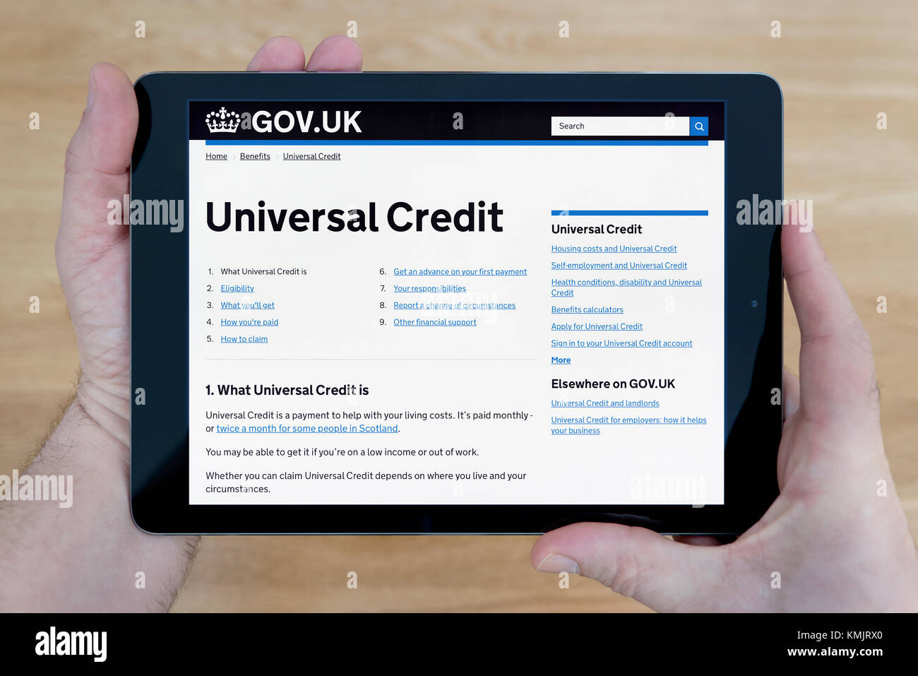 A man looks at the Universal Credit section of the Gov.uk website on an iPad tablet device, shot against a wooden table top background -Editorial only Stock Photo