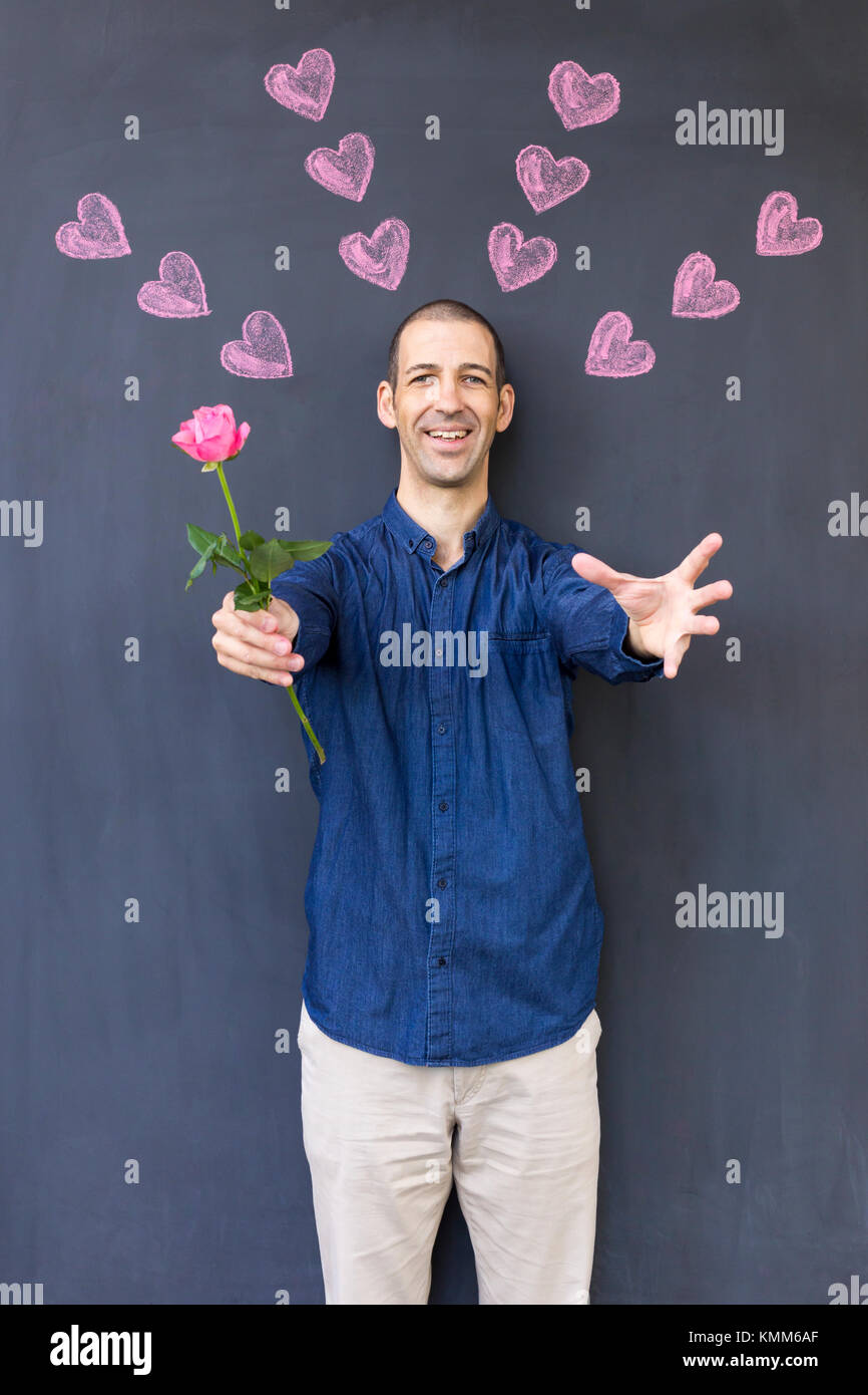 Single adult white man wearing a blue shirt standing in front of a blackboard with painted hearts holding a rose. Concept of crazy love. Stock Photo