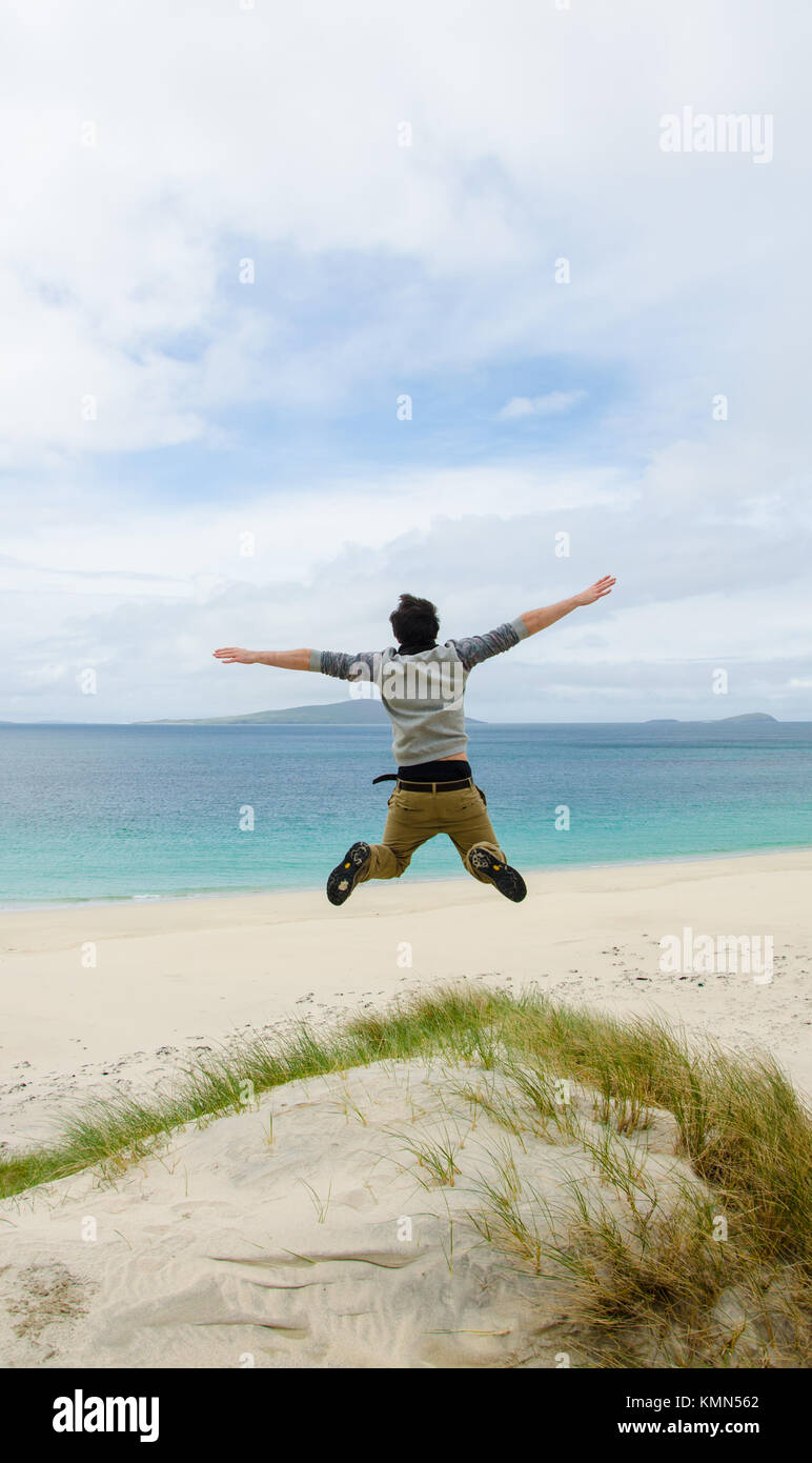 Young model jumping on a sand dune with open arms. White sandy beach and blue sky in the background. Scotland, UK. Stock Photo