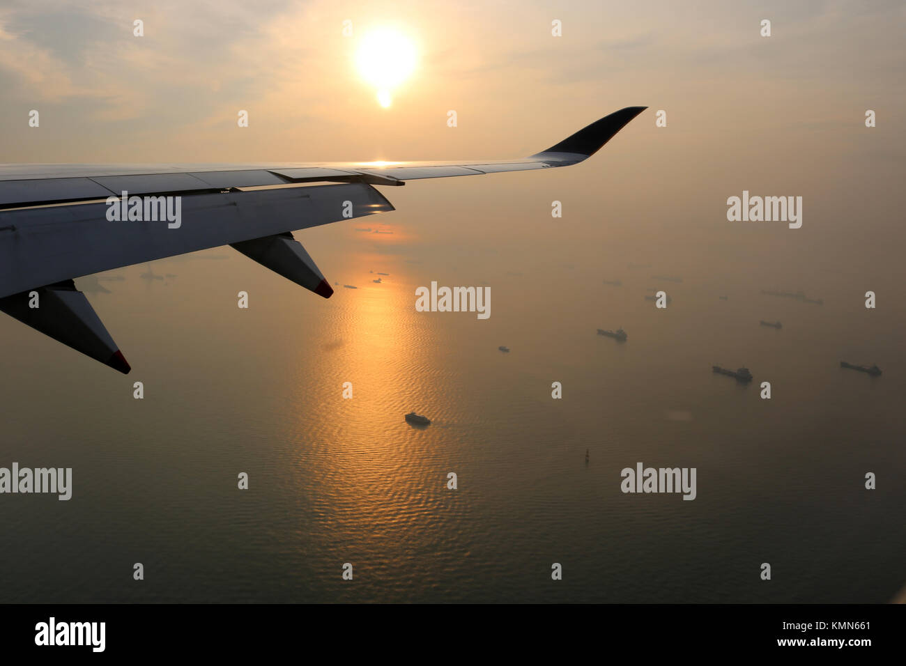 View from airplane of airplane wing over sunrise over Singapore strait with ships anchored in the water below. Sunrise also reflected off the water. Stock Photo