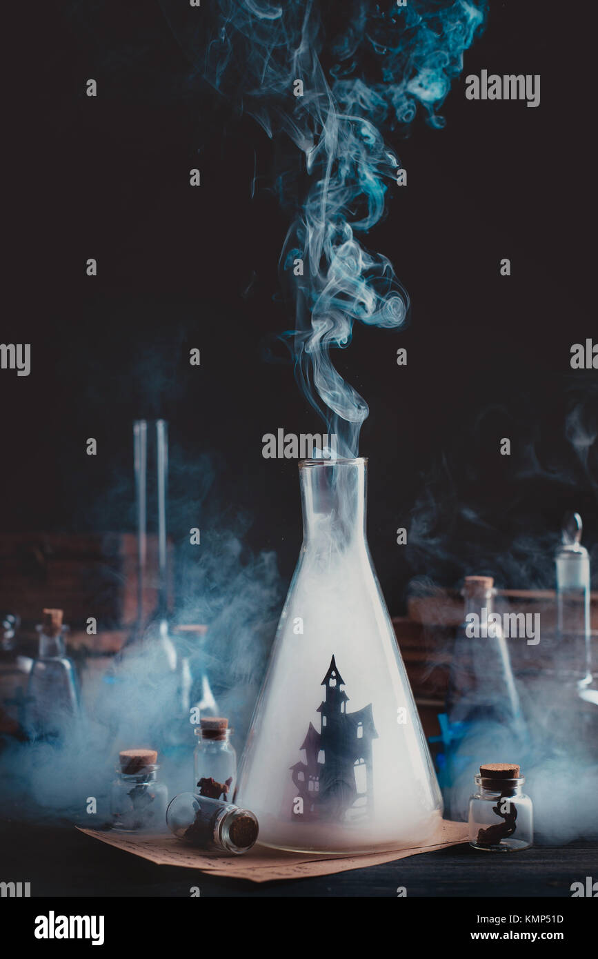 Lab bottle with haunted castle silhouette. Wizard workplace with smoke and magical equipment. Dark conceptual still life. Stock Photo