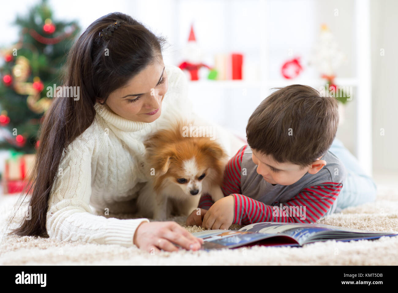 Merry Christmas and Happy New Year. Mom reading a book to her cute son near Christmas tree at home. Stock Photo