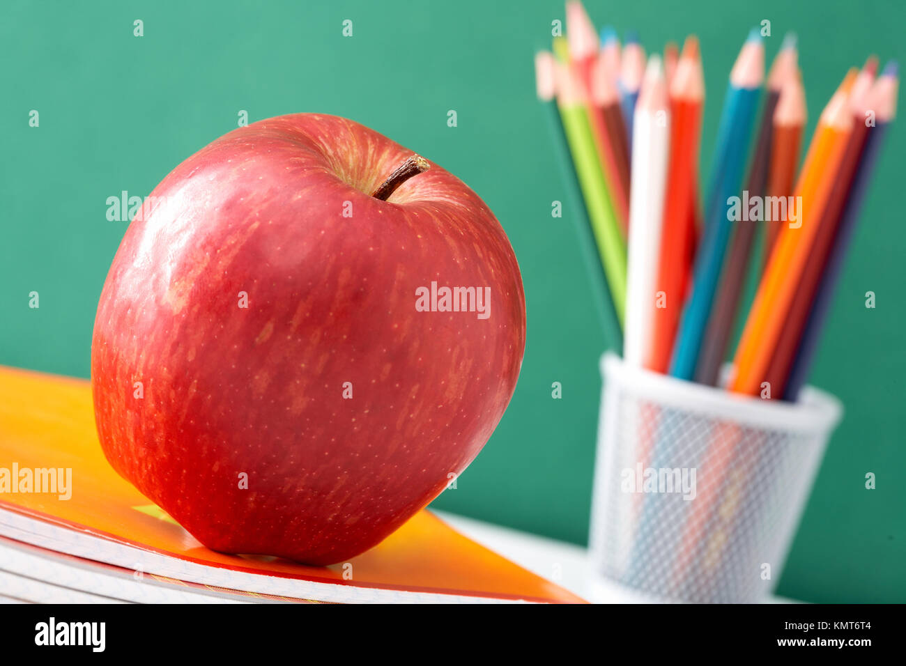 Close-up of big red apple on stack of copybooks with colorful pencils on background Stock Photo