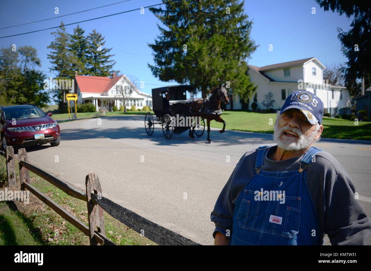 Old man in blue dungarees and baseball cap standing outside his house as an Amish buggy goes by in Charm, Ohio, USA Stock Photo