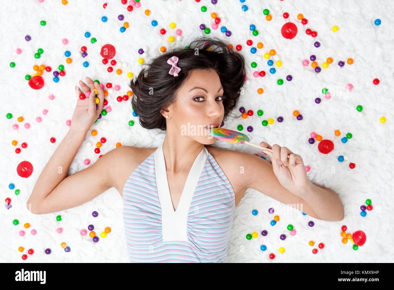 Young Latina woman laying on ruffled cloud like floor between colorful bubblegum balls eating a lollipop Stock Photo