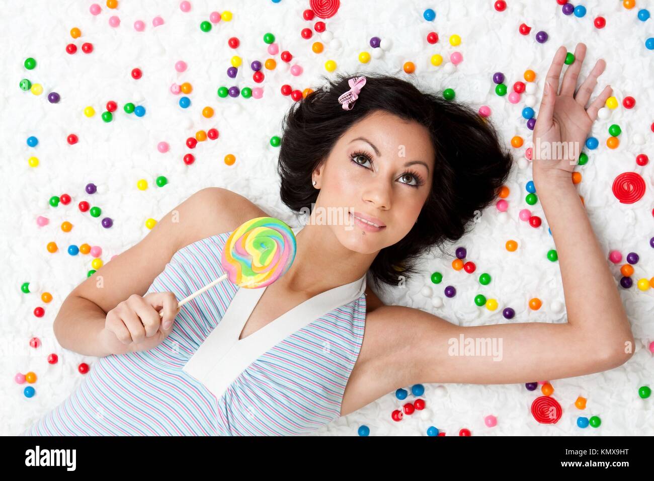 Young Latina woman laying on ruffled cloud like floor between colorful bubblegum balls holding a lollipop Stock Photo