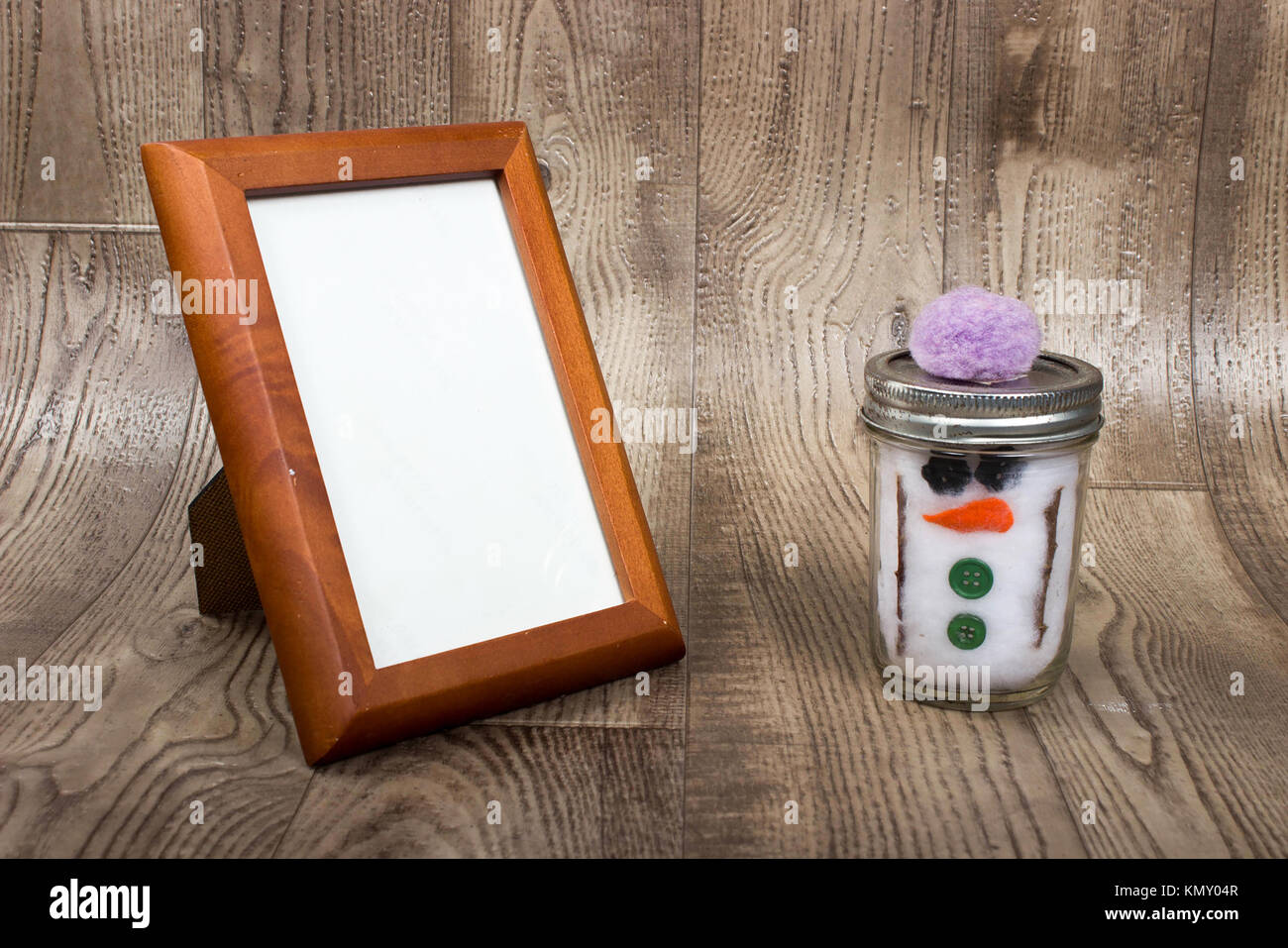 background of empty wooden  picture frame setting a wooden shelf with a hand crafted snowman made with twigs, buttons, and cotton balls in a jar. Stock Photo