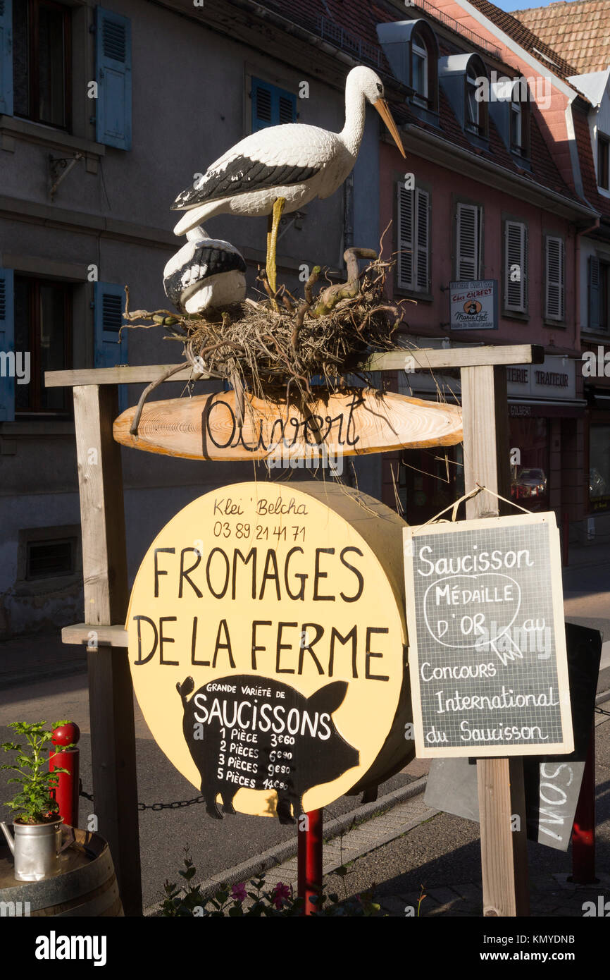 A sign outside a shop in Munster advertising local farm produced cheeses and sausages, adorned by a stork, regional symbol of Alsace Stock Photo