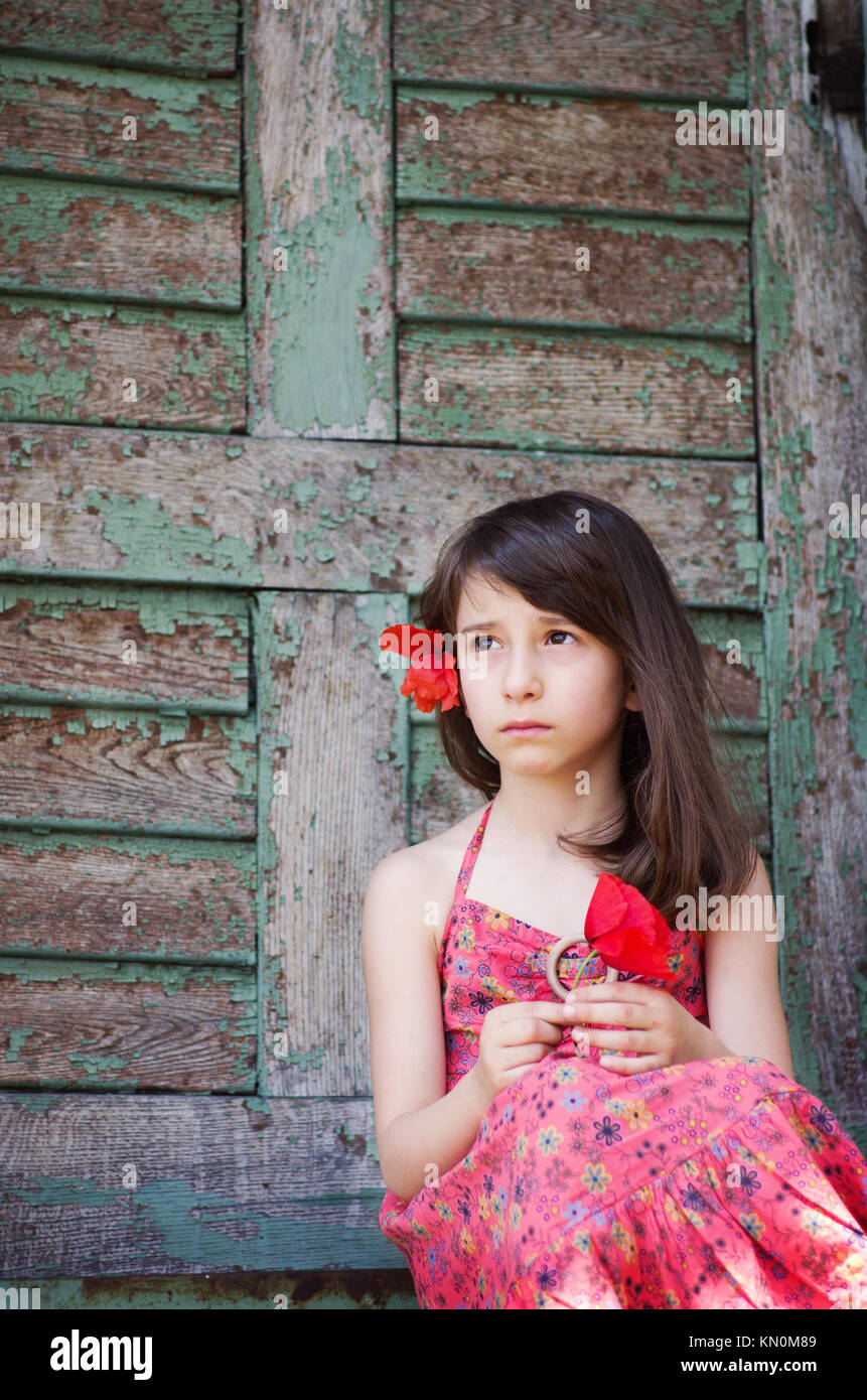 Pretty young girl 4-6 years sitting outside old wooden door and looking away. Poppy flower in her hands and in her hair. Dreamlike Stock Photo