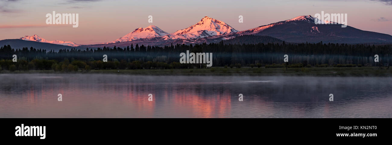 The Top of the Three Sisters Mountains and Mt. Bachelor turn Pink at Sunrise viewed from the Lake at Black Butte Ranch, Extra Wide Panorama Stock Photo