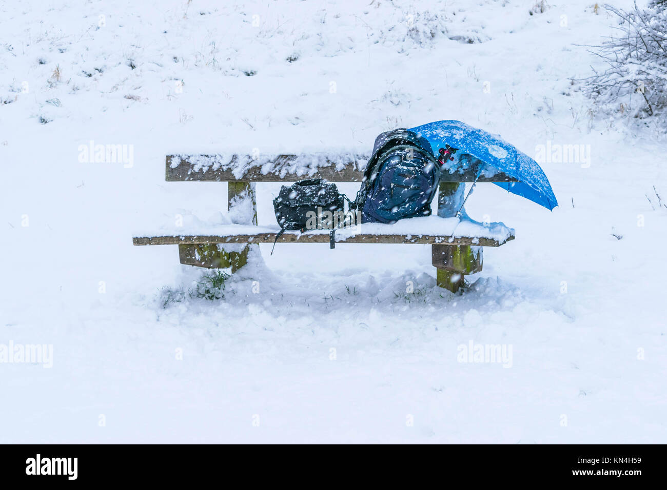 wooden bench chair in a park completely cover with snow after a blizzard during the winter christmas holidays, a backpack bag, umbrella, camera tripod and a small bag on the chair. Stock Photo