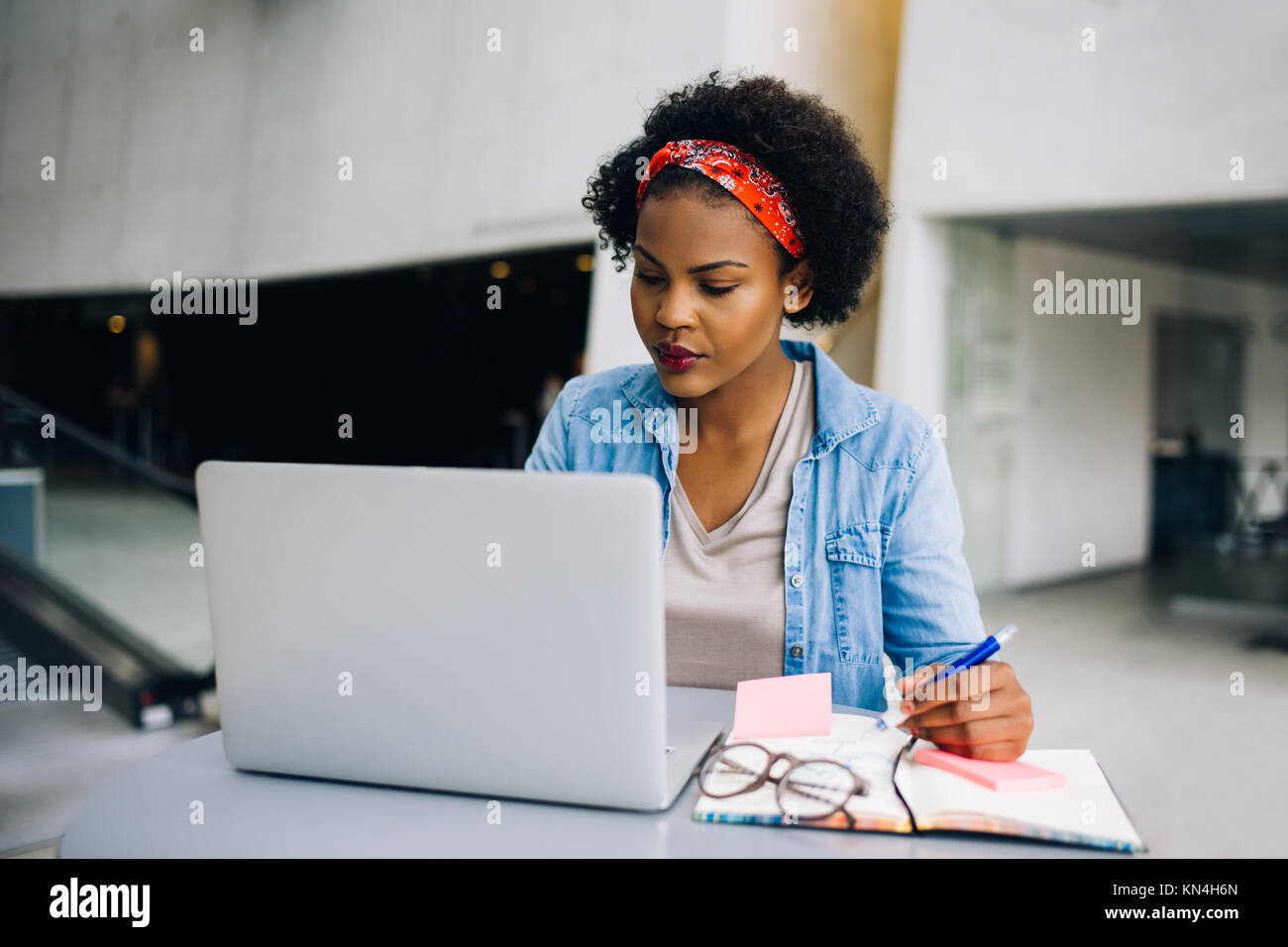 Young African female entrepreneur sitting at a table in a modern office building lobby working on a laptop and writing notes in her planner Stock Photo