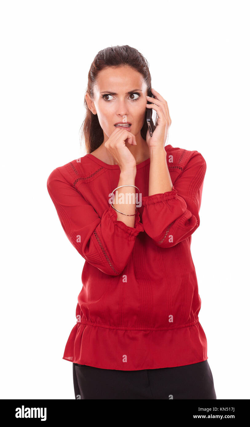 Portrait of angry hispanic lady on red shirt talking on her cell while standing on isolated white background - copyspace. Stock Photo