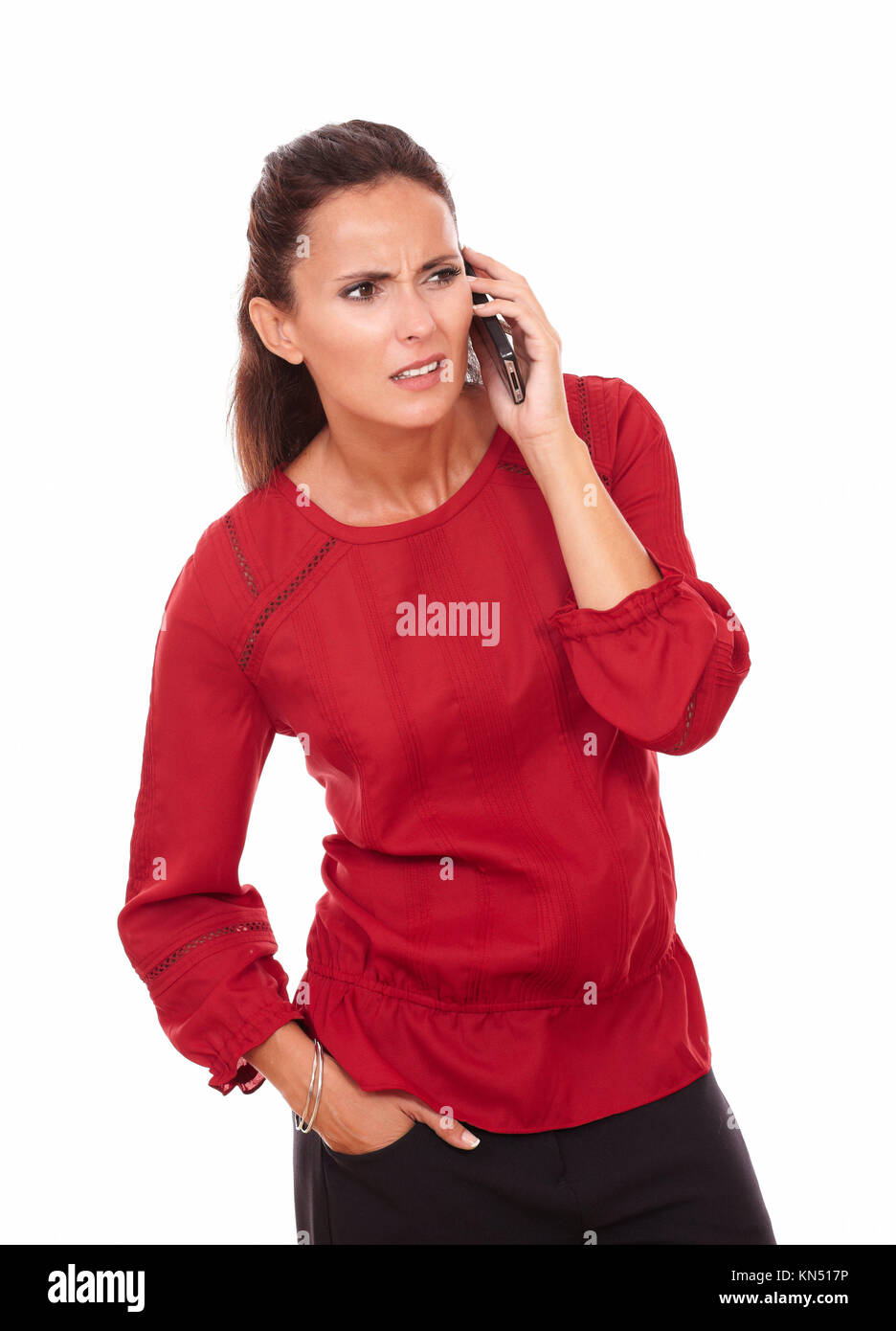 Portrait of confused hispanic lady on red blouse talking on her phone while looking to her left and standing on isolated white background - copyspace. Stock Photo