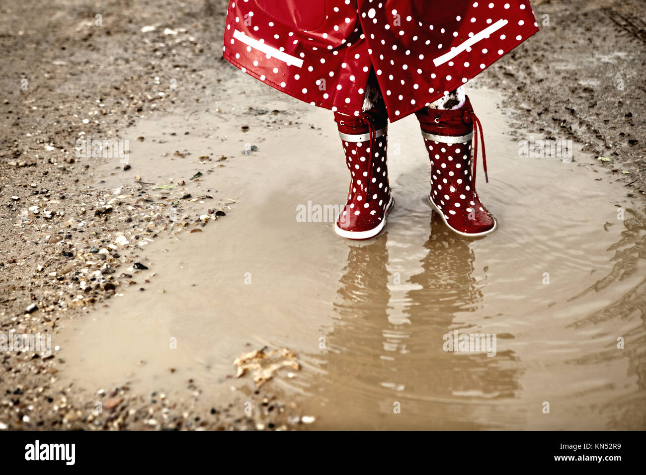 Detail of rainboots of a baby girl dressed with dotted raincoat in the puddles. Stock Photo